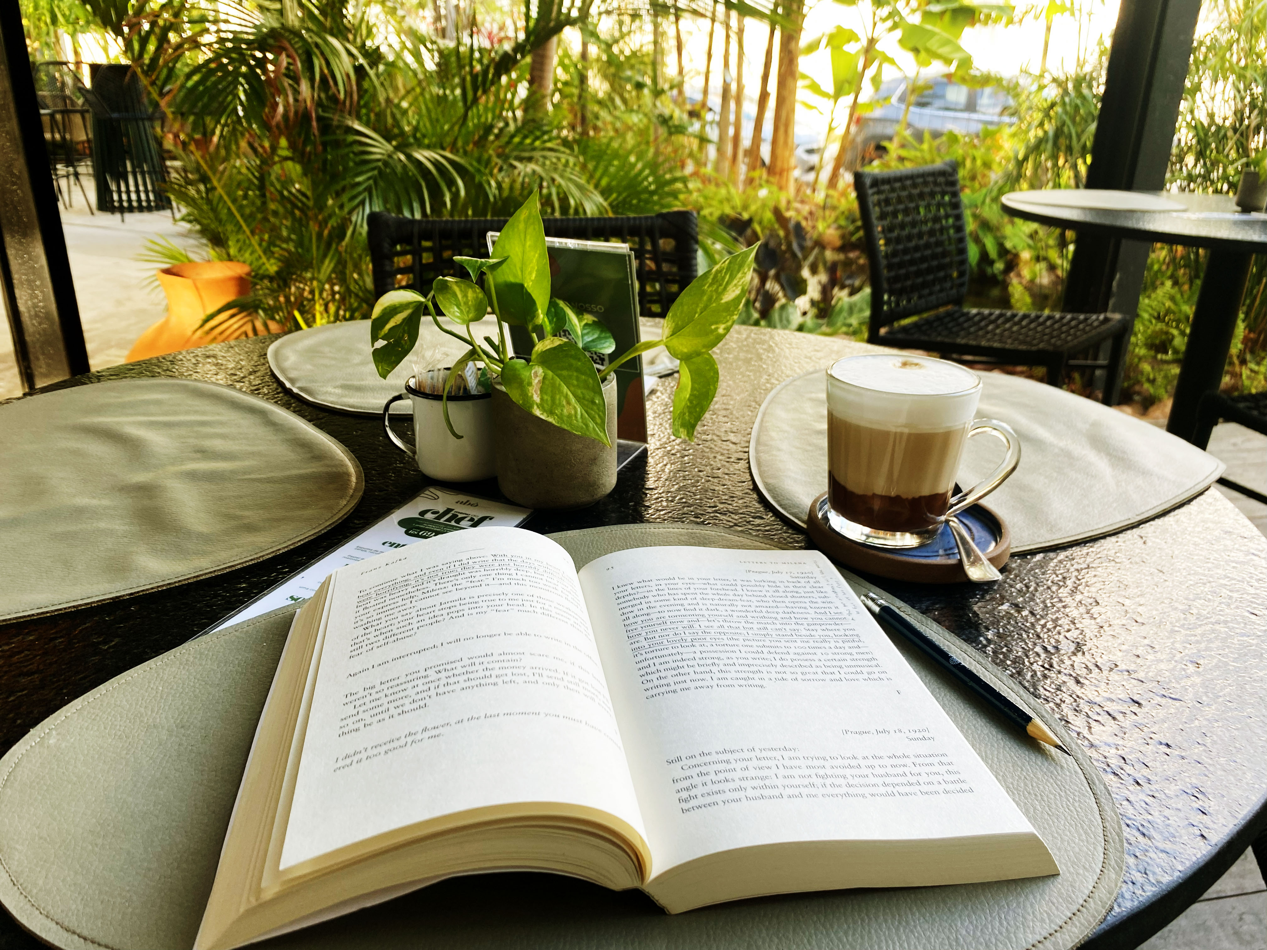 Em primeiro plano, um livro aberto com um marcador de páginas sobre uma mesa de vidro. Ao redor, há uma caneca de vidro transparente com café, um vaso pequeno com uma planta, pratos e talheres. O ambiente é um espaço externo, com a vegetação tropical e cadeiras desfocadas ao fundo, sob a luz do sol.